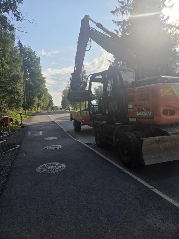 Gravemaskin på hjul, med henger, inntil fortauet på en vei